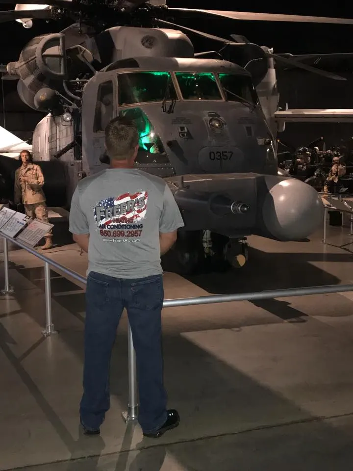 Mike Freer, seen from behind in a Freer's Heating & Air shirt, standing at the rail in front of an MH-53 PAVE LOW helicopter in a museum hangar